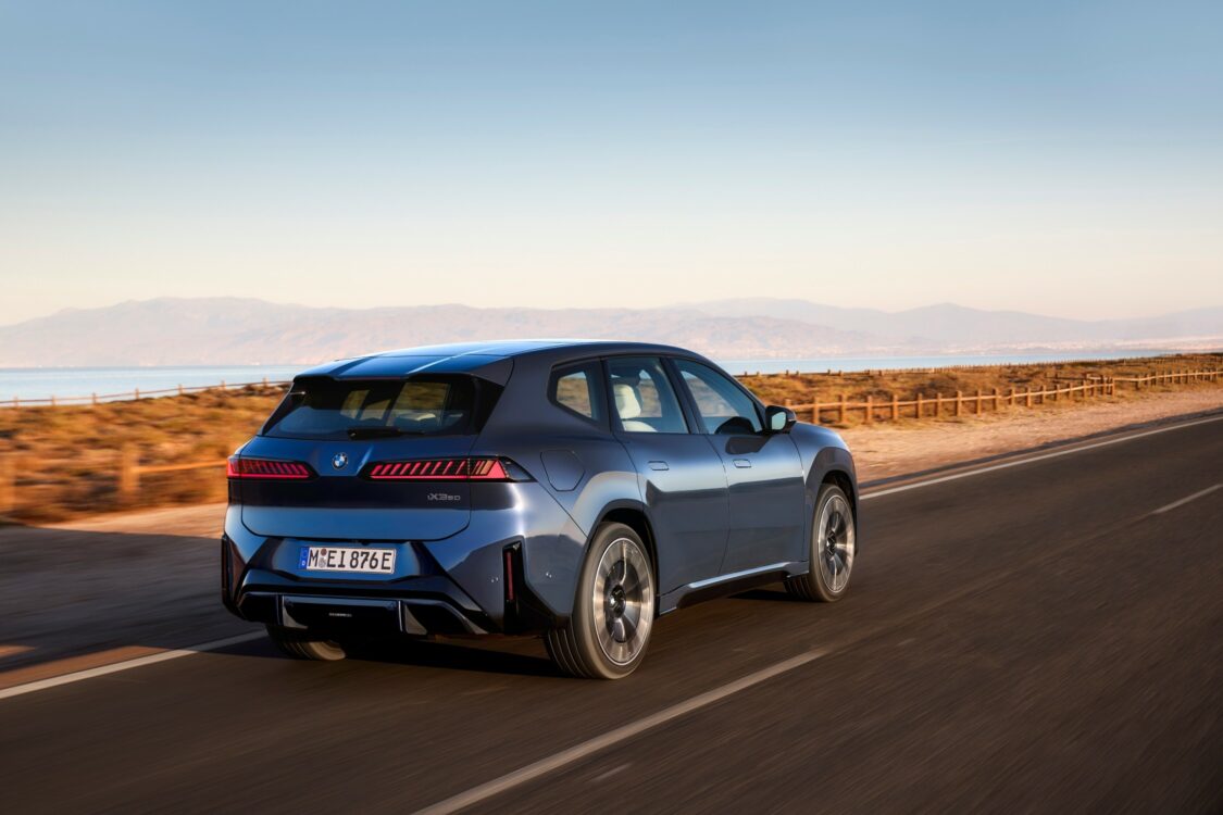 Vue arrière de la BMW iX3 roulant sur une route côtière, avec les feux arrière allumés et un paysage montagneux en arrière-plan.