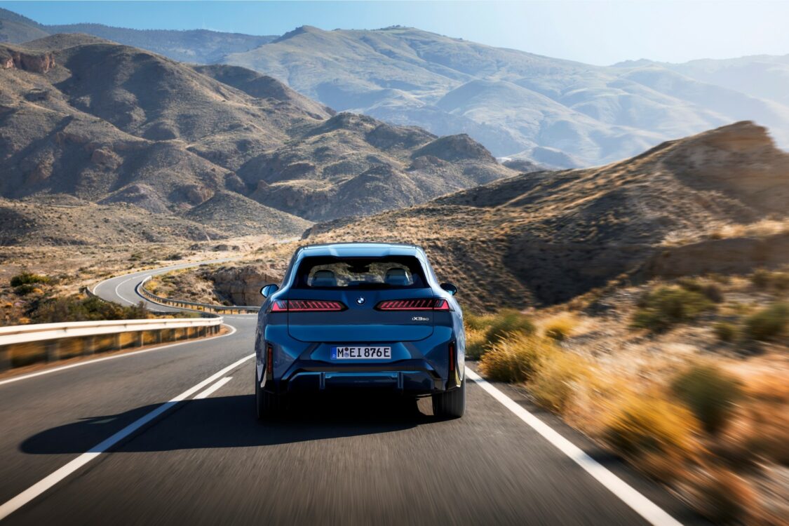 Vue arrière de la BMW iX3 roulant sur une route de montagne sinueuse, intégrée dans un paysage minéral et vallonné.