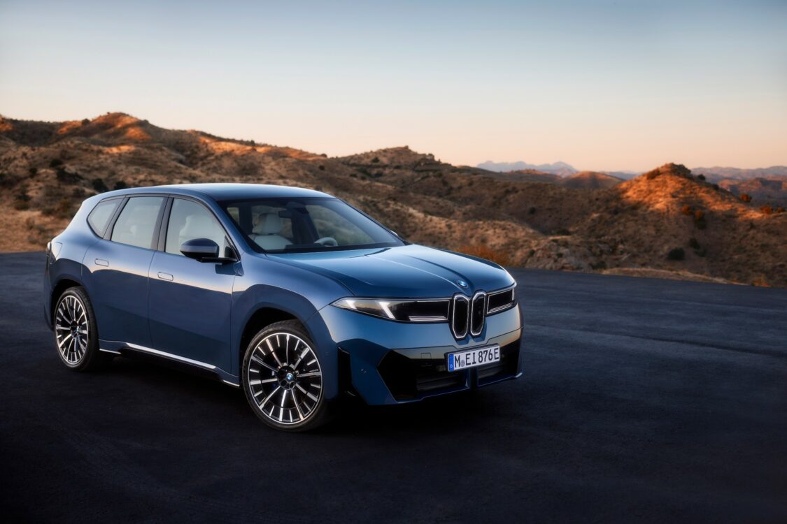 BMW iX3 bleue en vue avant trois quarts, stationnée sur une esplanade avec en arrière-plan un paysage montagneux au coucher du soleil.