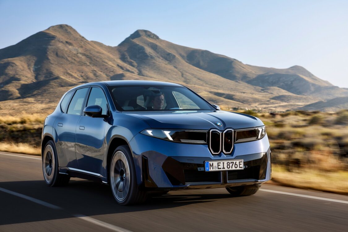 Vue extérieure de la BMW iX3 en position statique, captée sous un angle avant trois quarts. La carrosserie bleu métallisé se détache sur un décor de montagnes au soleil couchant, soulignant la modernité et la robustesse du SUV électrique. La face avant exprime puissance et raffinement, avec la calandre fermée et les projecteurs effilés caractéristiques de l’identité visuelle BMW.