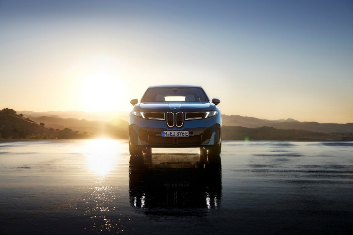BMW iX3 vue de face, stationnée sur une surface réfléchissante au coucher du soleil, avec les montagnes en arrière-plan.