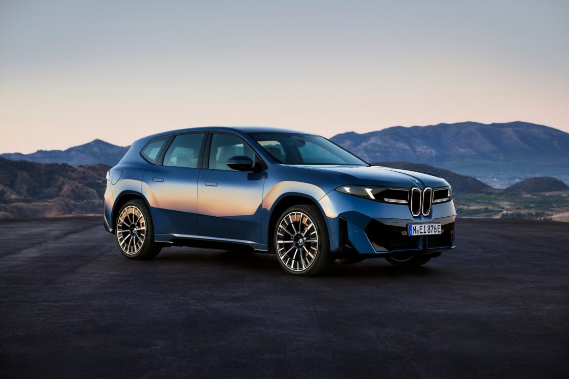Photographie frontale de la BMW iX3 bleue, immobile sur une surface humide qui reflète ses projecteurs et sa calandre double emblématique. L’arrière-plan dévoile un paysage montagneux baigné par la lumière chaude du coucher de soleil. Cette composition renforce l’identité visuelle du SUV électrique, associant puissance et élégance dans un cadre presque cinématographique.