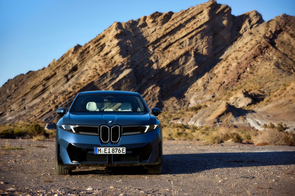 BMW iX3 vue de face, carrosserie bleue, stationnée devant une formation rocheuse inclinée dans un paysage désertique.