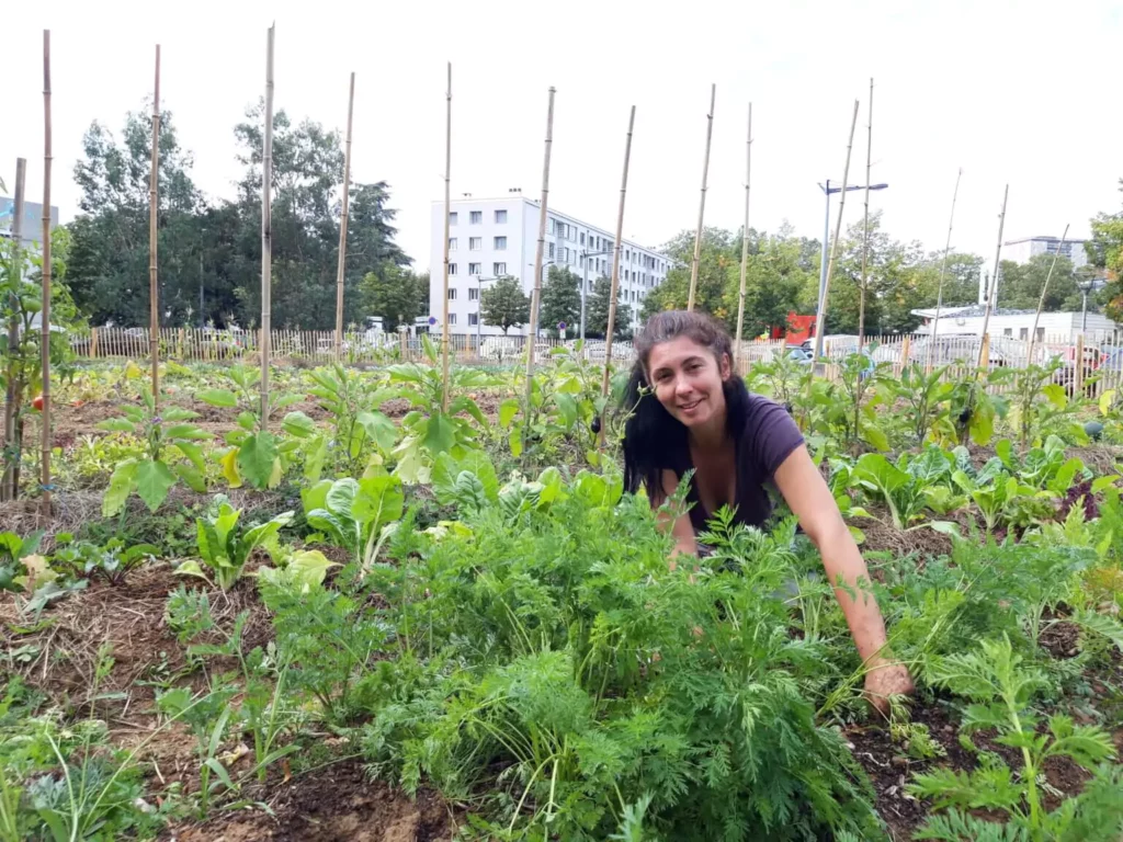 Femme souriante récoltant des carottes dans une ferme urbaine en pleine terre, au cœur d’un quartier résidentiel à Lyon-La Duchère