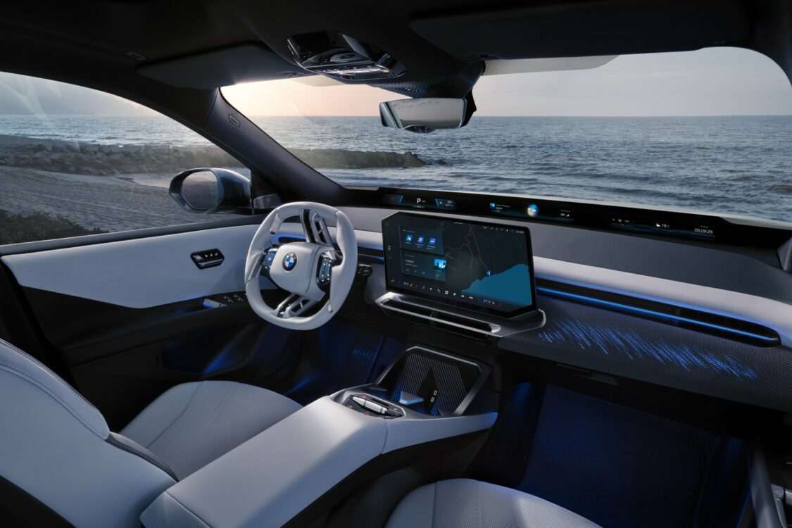 Vue intérieure de la BMW iX3 côté conducteur, avec volant blanc, écran central affichant la navigation et décor LED bleuté face à un paysage marin