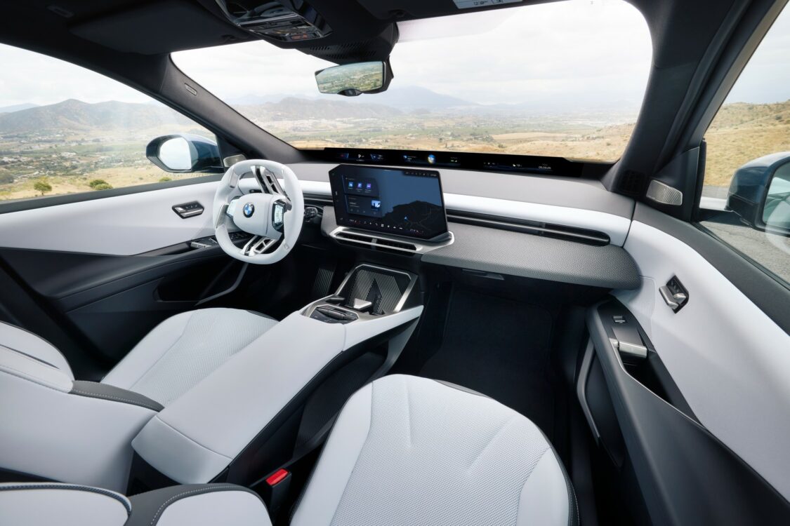 Intérieur de la BMW iX3 vu côté passager, avec sièges blancs, volant blanc et grand écran central, sur fond de paysage montagneux.