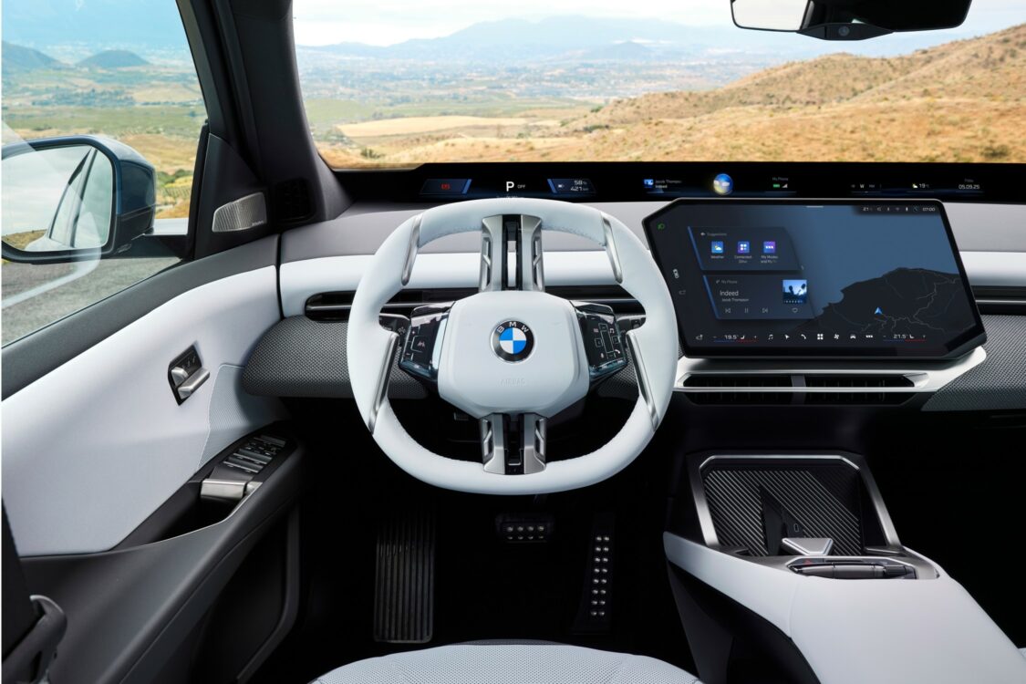 Gros plan sur le volant blanc et l’écran central de la BMW iX3, avec affichage de navigation, sur fond de paysage montagneux.