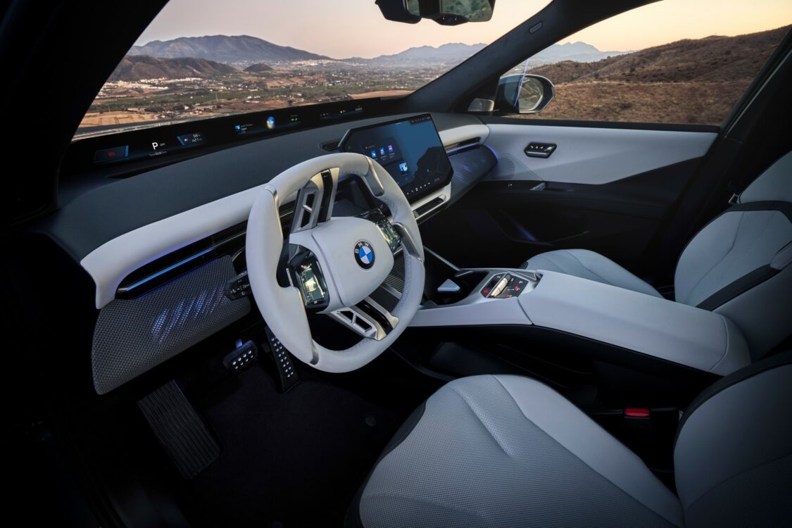 Vue oblique de l’intérieur de la BMW iX3, montrant le volant blanc, l’écran central et la console, avec un paysage montagneux en arrière-plan.